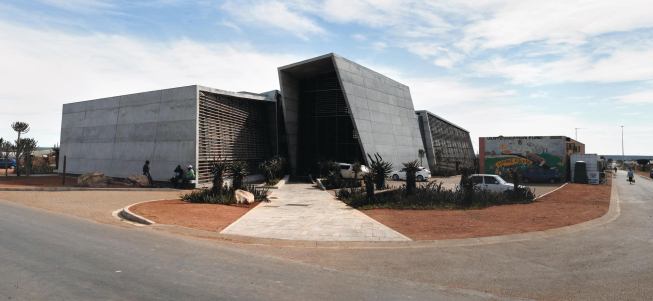 The concrete, glass, and gum-pole-clad volumes of the Ubuntu Centre stand out amid the urban fabric of Zwide, but the massing is intentionally broken up into multiple volumes to avoid overwhelming neighboring buildings. On the south façade, the education wing anchors the corner, followed by the community hall and clinic.