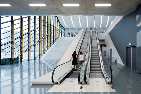 On the ground floor, visitors enter under the cantilevered form of the médiatheque and use escalators to access the upper levels. Floor detailing echoes the lines of the louvers and the lights above.